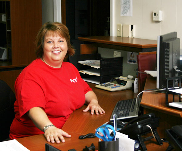 Woman in red t shirt smiling at camera and sitting at a desk